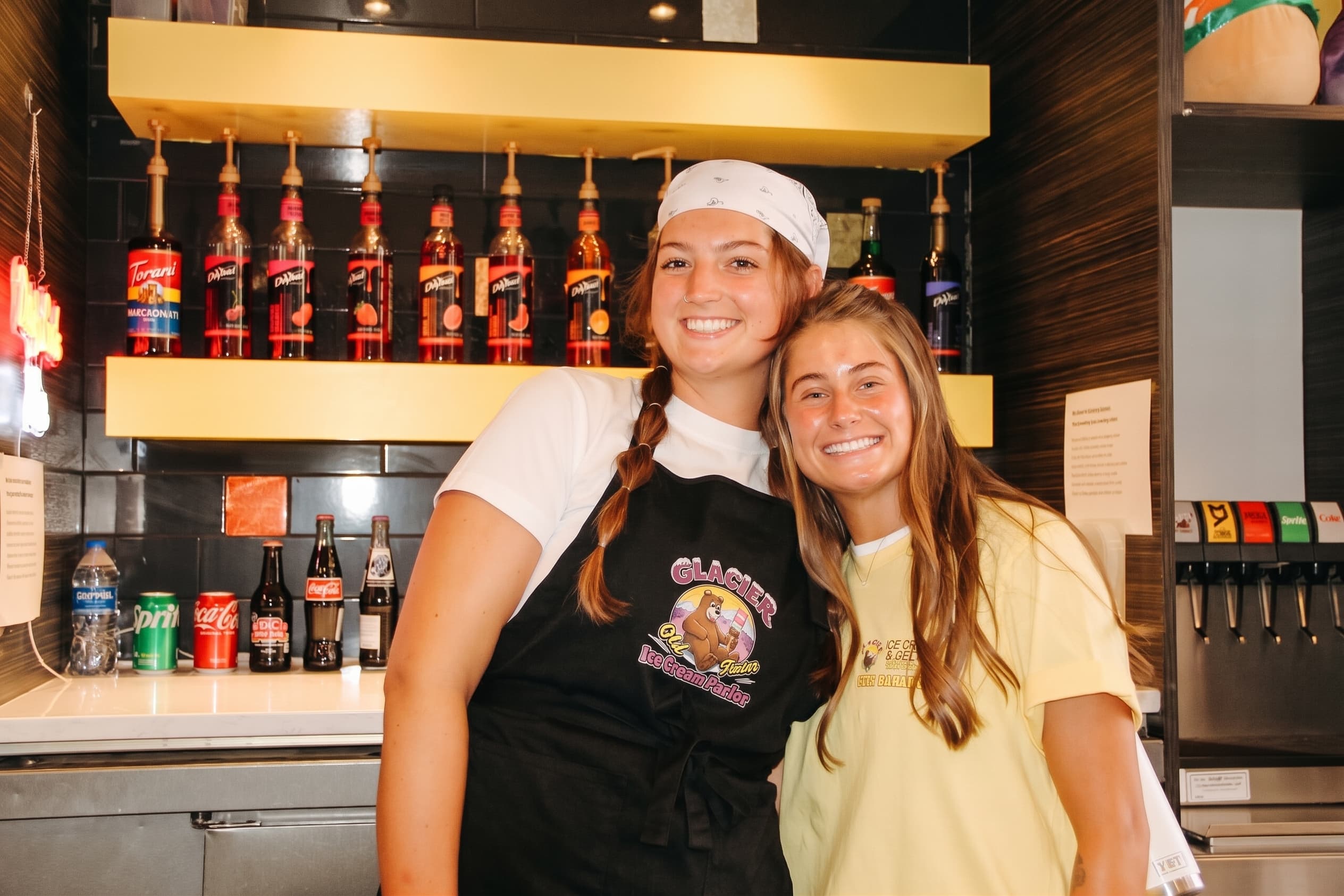 Two crew members behind the soda counter under the syrup shelf